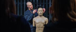A workshop host in dark sweater putting on headphones on a mannequin.